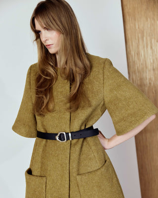 Woman wearing a long mustard yellow coat with a belt against a white background with a leather belt