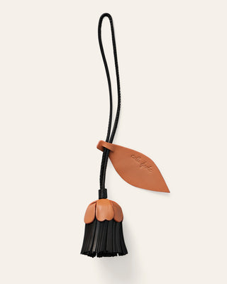 A small leather accessory with a flower strap and leaf-shaped tab, attached to a dangling flower shaped charm in black and natural beige, shown on a plain light warm-gray background.