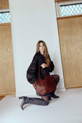 Cala Jade humi - Woman holding a brown leather bag against a white wall with wooden panels.