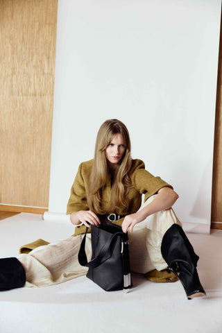 Woman sitting on the floor holding a black handbag against a white wall.