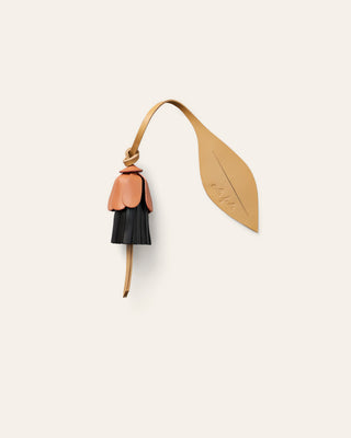 A small leather accessory with a flower beige-brown strap and leaf-shaped tab, attached to a dangling bell-shaped charm in red and natural beige, shown on a plain light warm-gray background.