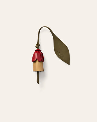 A small leather accessory with a flower olive-brown strap and leaf-shaped tab, attached to a dangling bell-shaped charm in red and natural beige, shown on a plain light warm-gray background.