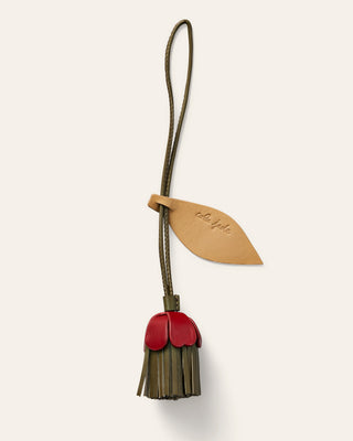 A small leather accessory with a flower  strap and leaf-shaped tab, attached to a dangling flower shaped charm in red and natural beige, shown on a plain light beige background.