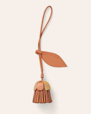 A small leather accessory with a flower  strap and leaf-shaped tab, attached to a dangling flower shaped charm in red and natural beige, shown on a plain light warm-gray background.