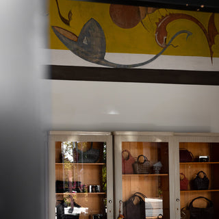 A retail interior showing two tall glass-front display cabinets filled with handbags and accessories, with a table in the foreground and a decorative mural on the wall above.