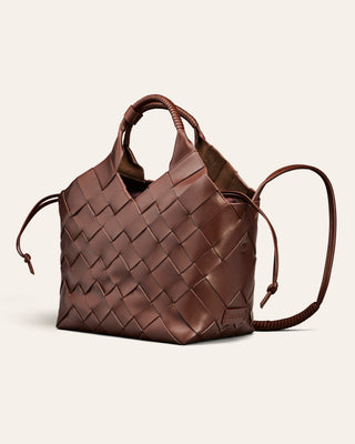 A brown woven leather handbag with integrated top handles and a long shoulder strap, photographed on a clean off-white background.