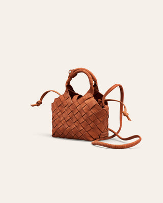 A small woven leather handbag in a warm rust-brown color, featuring a top handle, a long shoulder strap, and a knotted detail on the side, set against a solid off-white background.