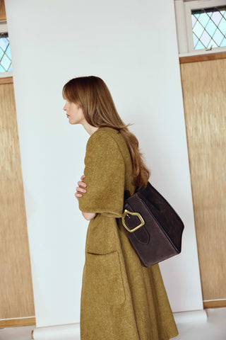 Woman in a long green coat holding a brown leather bag against a white wall with wooden panels.