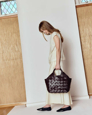 Woman holding a patterned handbag in a minimalistic room with wooden floors and large windows.