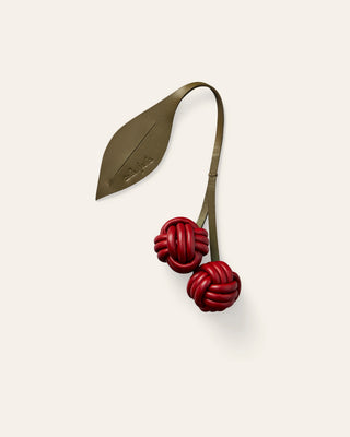A leather bag charm with red knotted cherries on a curved olive-green stem with a leaf, on a beige background.