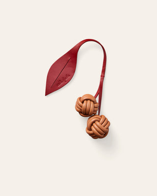 A leather bag charm with red knotted cherries on a curved red stem with a leaf, on a beige background.